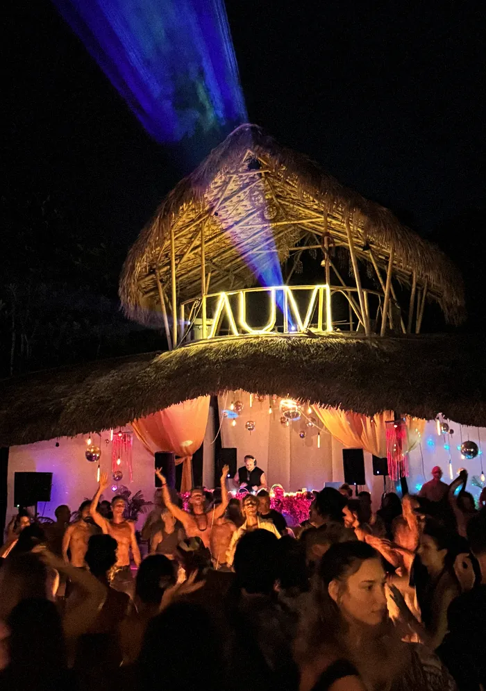Dancers in front of a thatched DJ tower lit with the word AUM, a beam of light cutting through the night sky