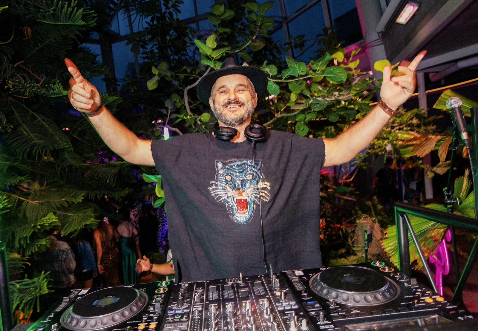 Tristan Gorski behind the decks in a black hat, grinning, both arms raised wide, surrounded by tropical plants