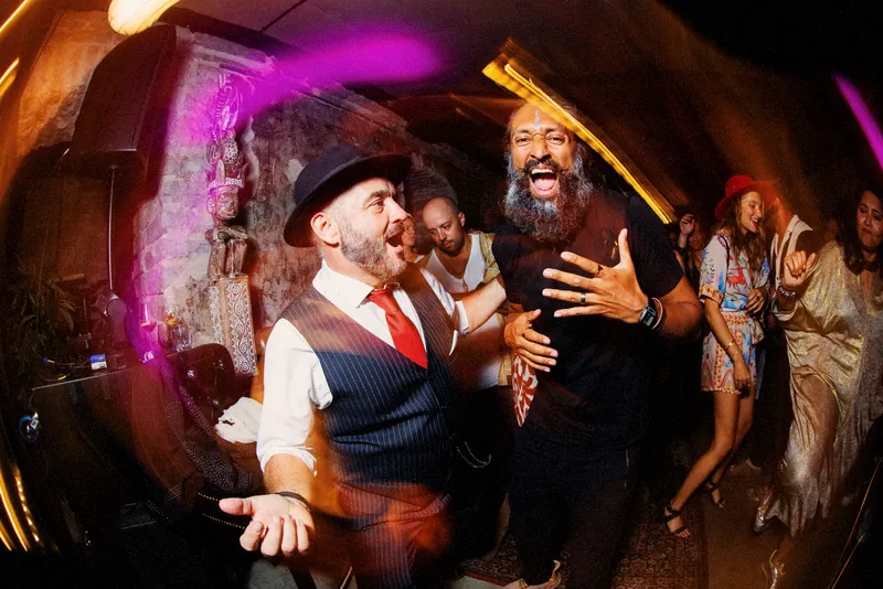Tristan Gorski in a hat and red tie, roaring with laughter alongside a bearded friend on a crowded dance floor
