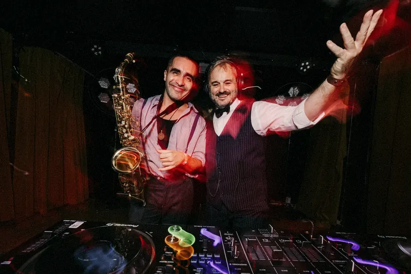 Tristan Gorski behind the decks in a waistcoat and bow tie, one arm flung out, next to a smiling saxophone player