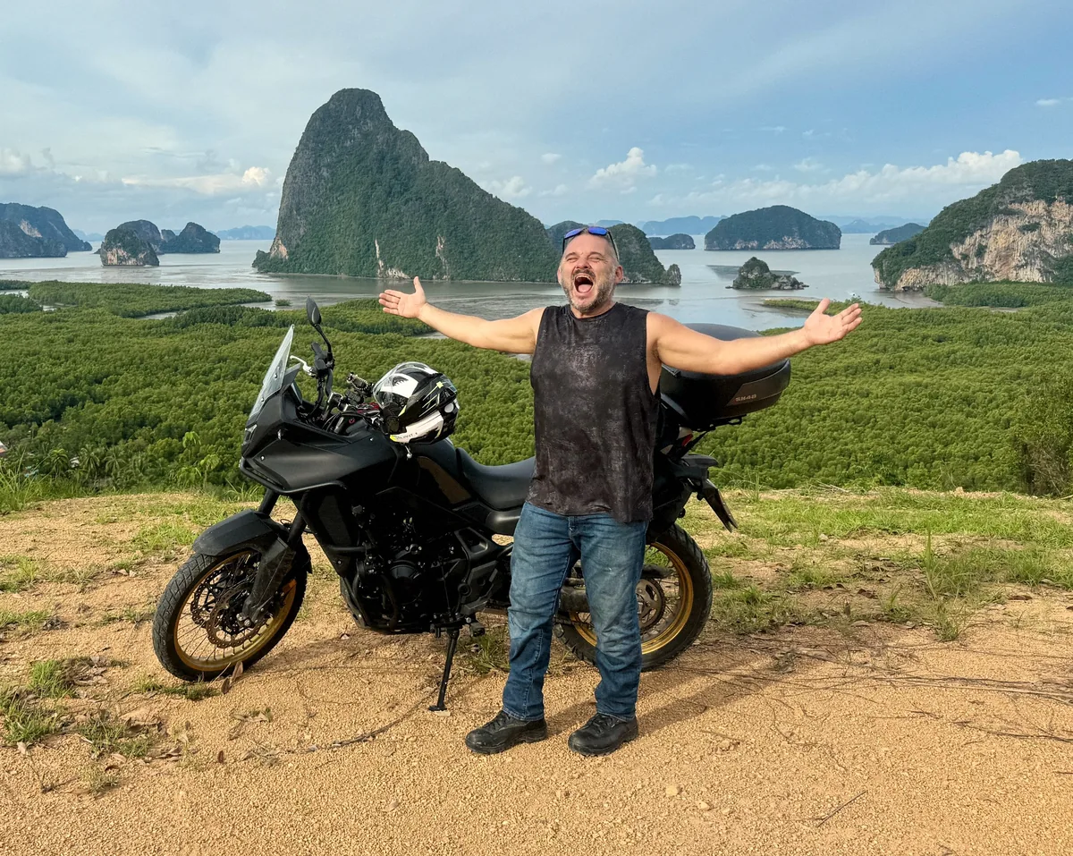 Tristan Gorski standing in front of his adventure motorbike with his arms flung wide, laughing, with limestone islands, sea and mangrove forest behind him